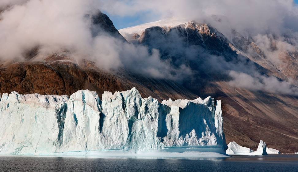 Gran glaciar flotando en Groenlandia frente a montañas áridas cubiertas parcialmente por nubes bajas.